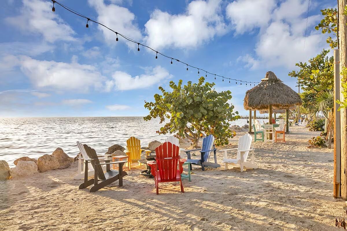 Une plage avec des chaises Adirondack aux couleurs vives, une structure à toit de chaume et une guirlande de lumières.