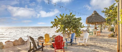 Une plage avec des chaises Adirondack aux couleurs vives, une structure à toit de chaume et une guirlande de lumières.