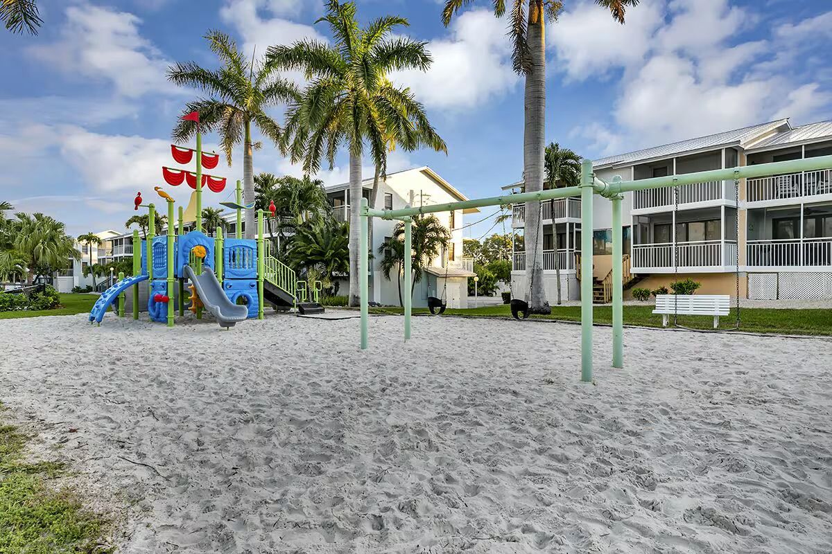 A playground with slides and climbing structures, surrounded by palm trees and a building with balconies.