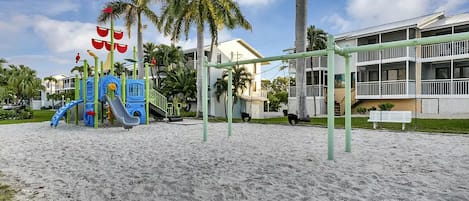 Un parque infantil con toboganes y estructuras para escalar, rodeado de palmeras y un edificio con balcones.