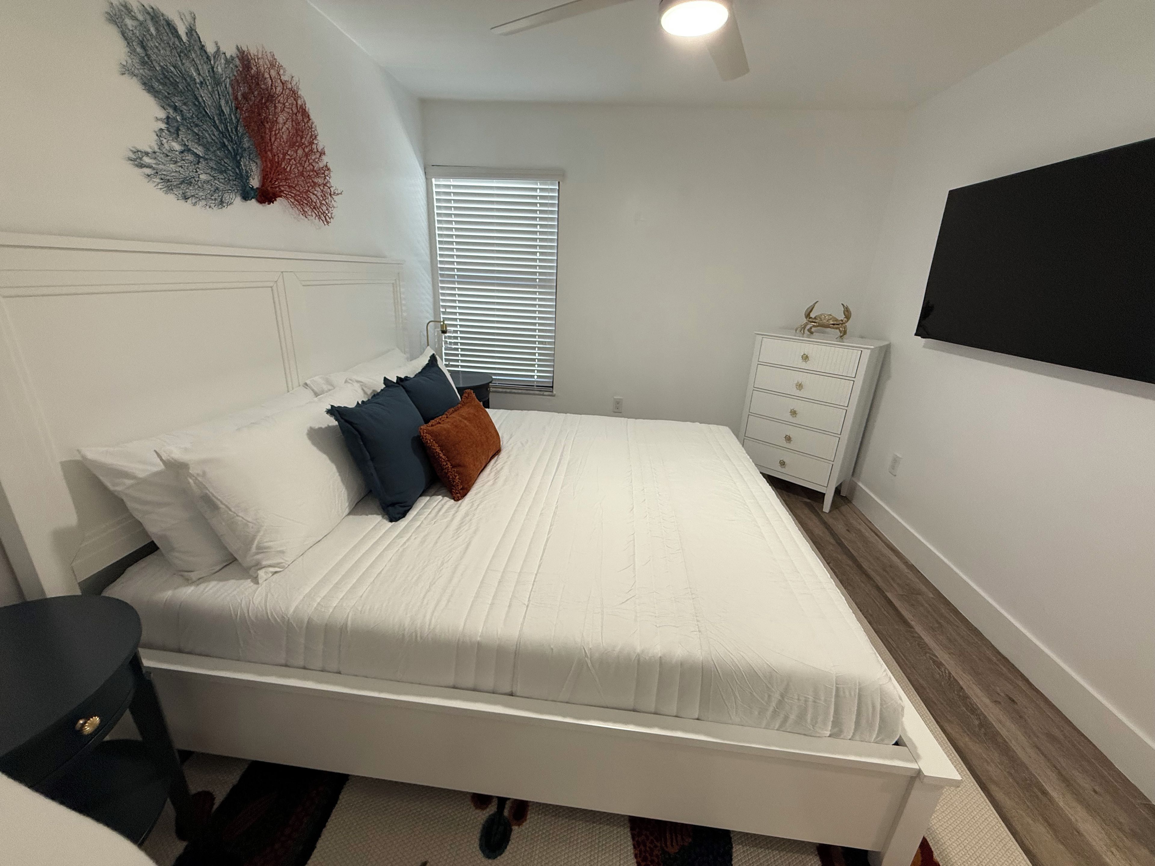A bedroom with a white bed, a dresser, a nightstand, and a window with blinds.