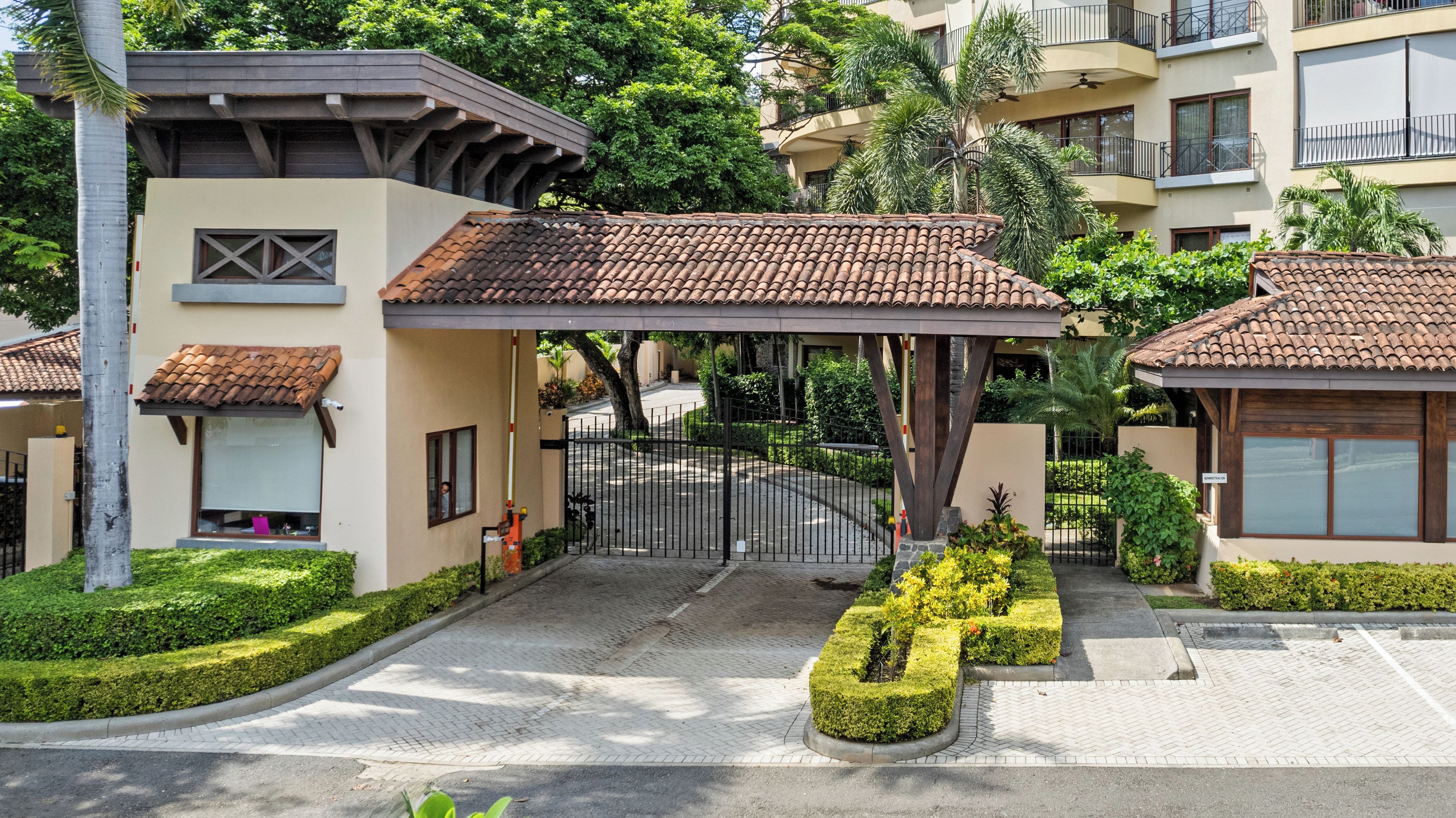 A gated entrance with a covered walkway, a well-maintained garden, and a multi-story building with balconies.
