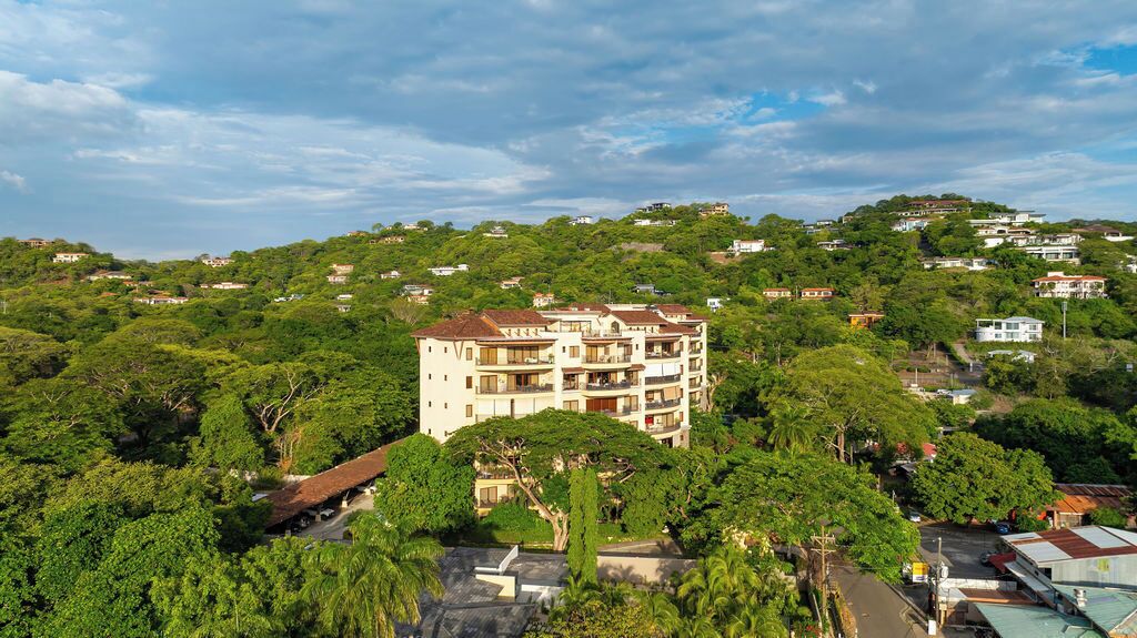 A multi-story building surrounded by dense greenery, with a clear sky above.