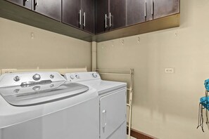A laundry room with a front-loading washer and dryer, wooden cabinets above, and a blue chair to the right. - Hermosa del Mar 1-6B | Pools & Gym, Walk to Beach (Playa Hermosa)