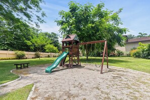 A playground with a slide and swings, surrounded by grass and trees. - Hermosa del Mar 1-6B | Pools & Gym, Walk to Beach (Playa Hermosa)