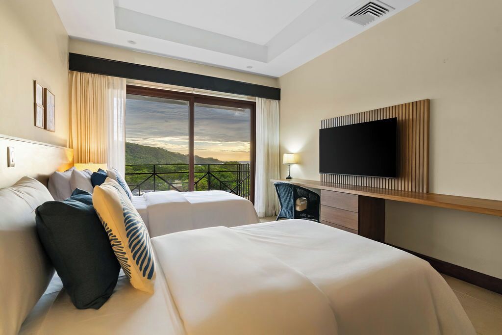 A hotel room with two beds, a TV, a desk, and a balcony with a view.