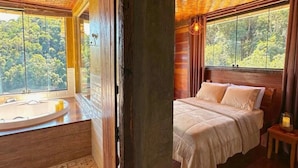 Ein Schlafzimmer mit einem großen Bett, einem Nachttisch, einem Fenster mit Blick auf den Wald und einer Badewanne mit Handtuch.