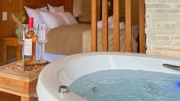 A bedroom with a large bathtub, a bedside table with a bottle and glasses, and a bed with pillows.