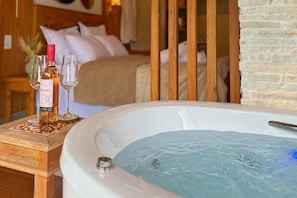 A bedroom with a large bathtub, a bedside table with a bottle and glasses, and a bed with pillows.