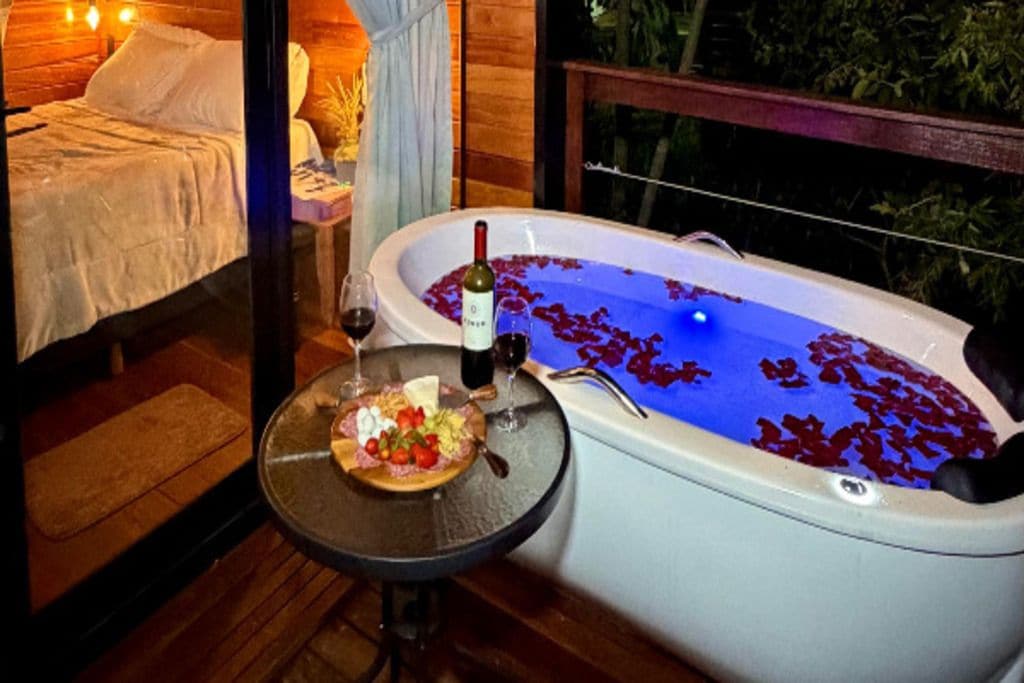 Quarto com cama, banheira cheia de água e pétalas de rosa, e mesa com prato de comida e copos de vinho.