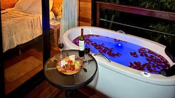 Quarto com cama, banheira cheia de água e pétalas de rosa, e mesa com prato de comida e copos de vinho.