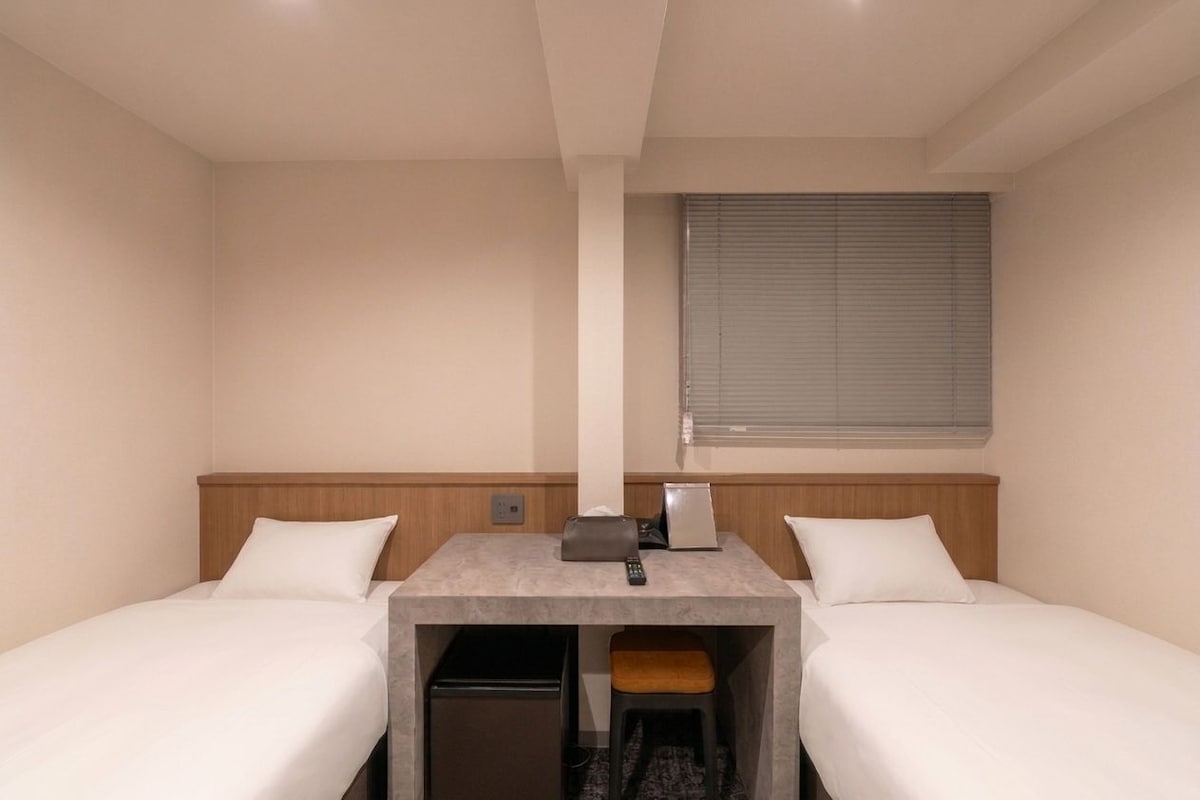 Una habitación de hotel con dos camas individuales, un escritorio pequeño con una computadora portátil, un control remoto y una ventana con persianas.