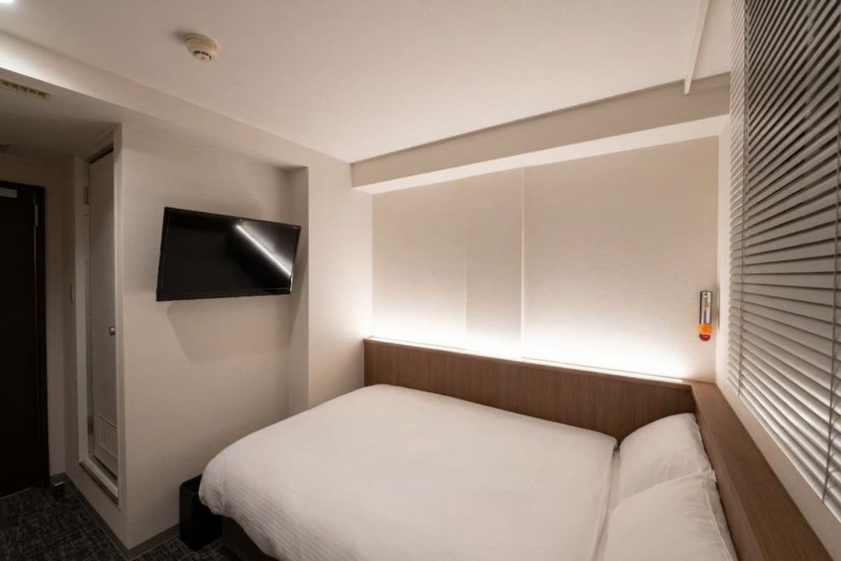Habitación de hotel con una cama, un televisor montado en la pared y una ventana con persianas.