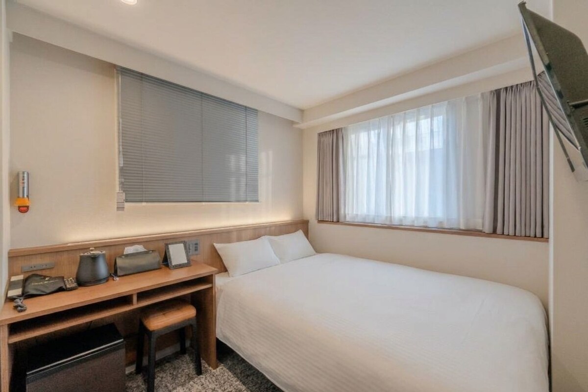 Una habitación de hotel con cama, escritorio con silla, televisor y ventana con cortinas.