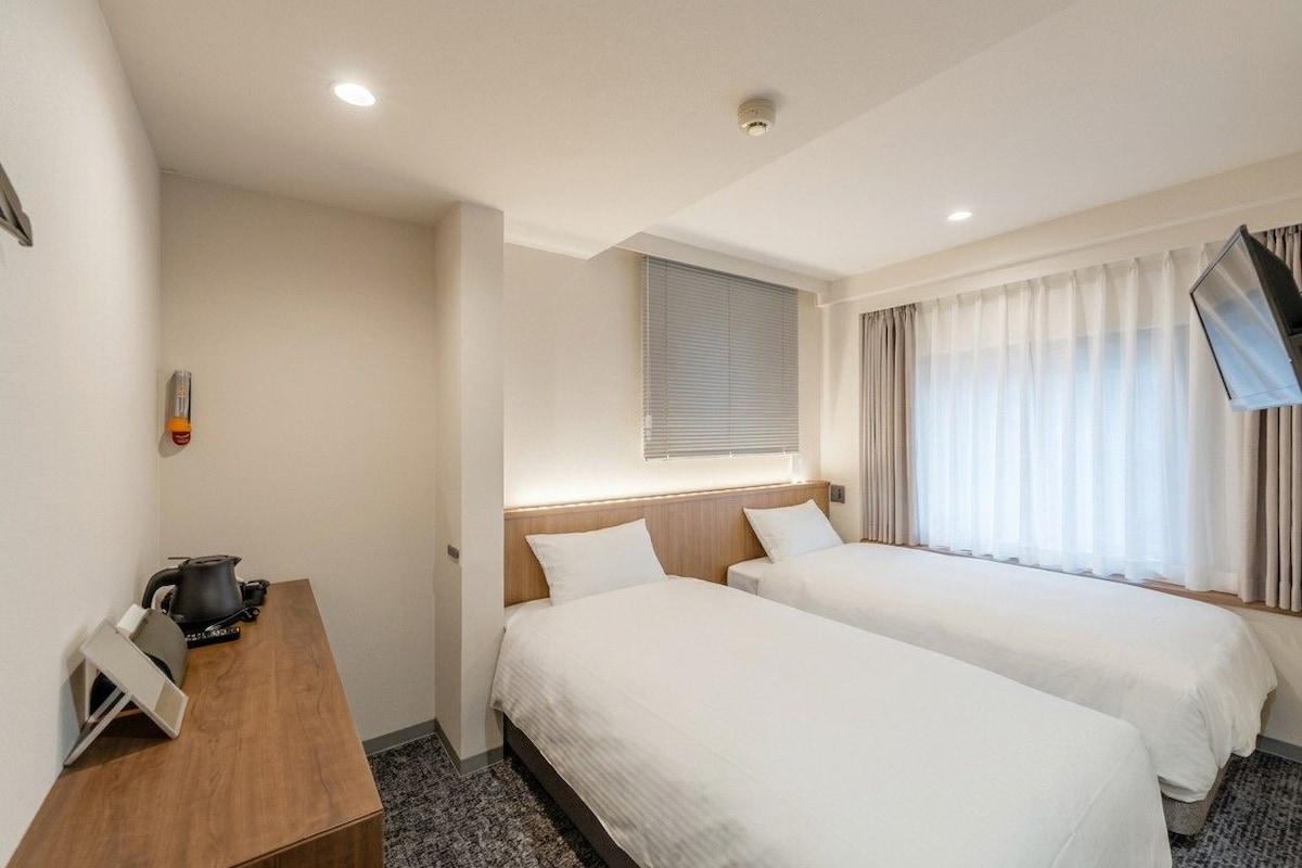 Habitación de hotel con dos camas, un escritorio con hervidor, televisión de pantalla plana y ventana con cortinas.