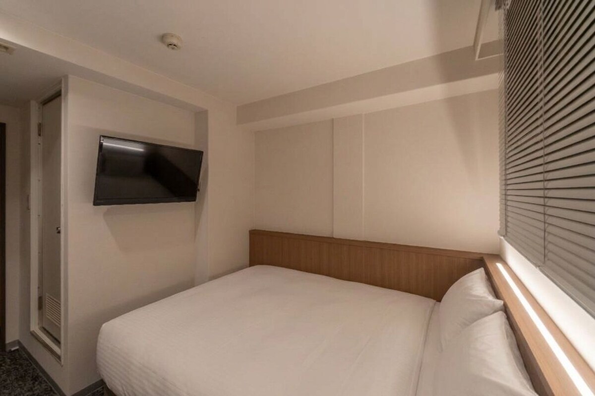 Una habitación de hotel con una cama, un televisor montado y una ventana con persianas.