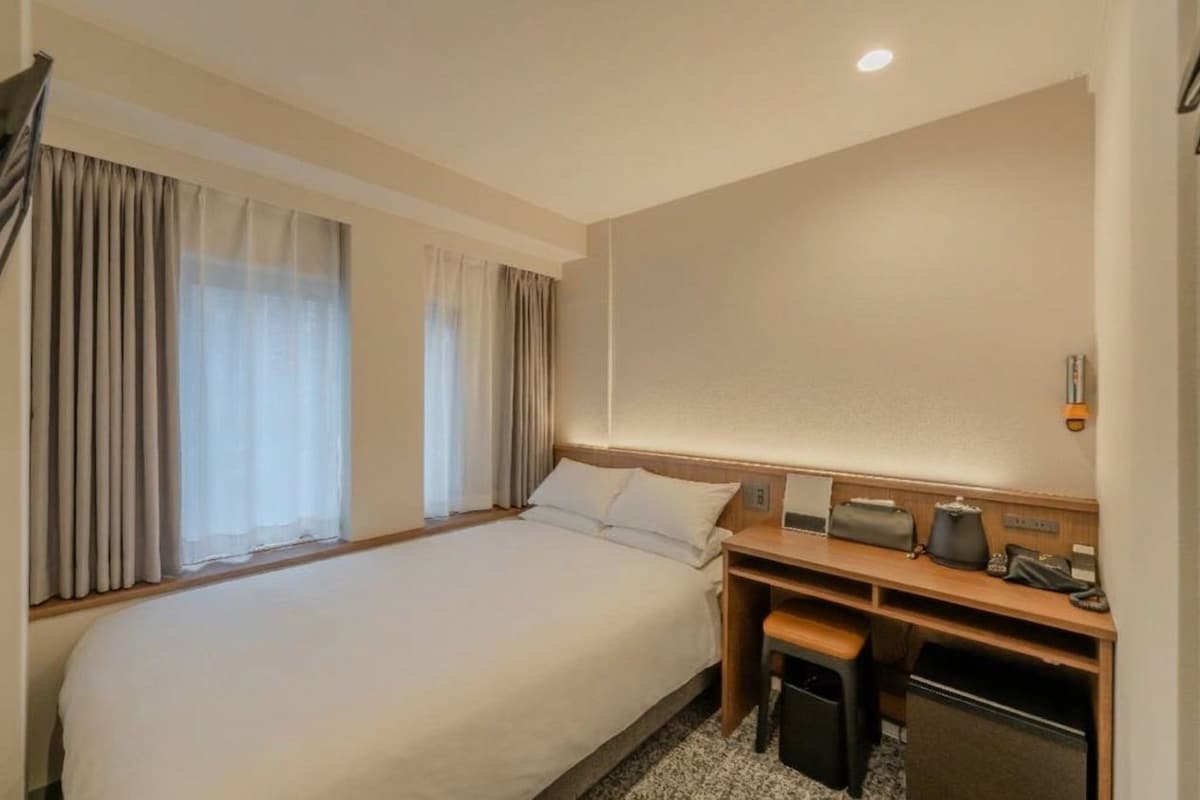 Una habitación de hotel con una cama grande, un escritorio con una silla, un bote de basura pequeño y una mesita de noche con una lámpara.