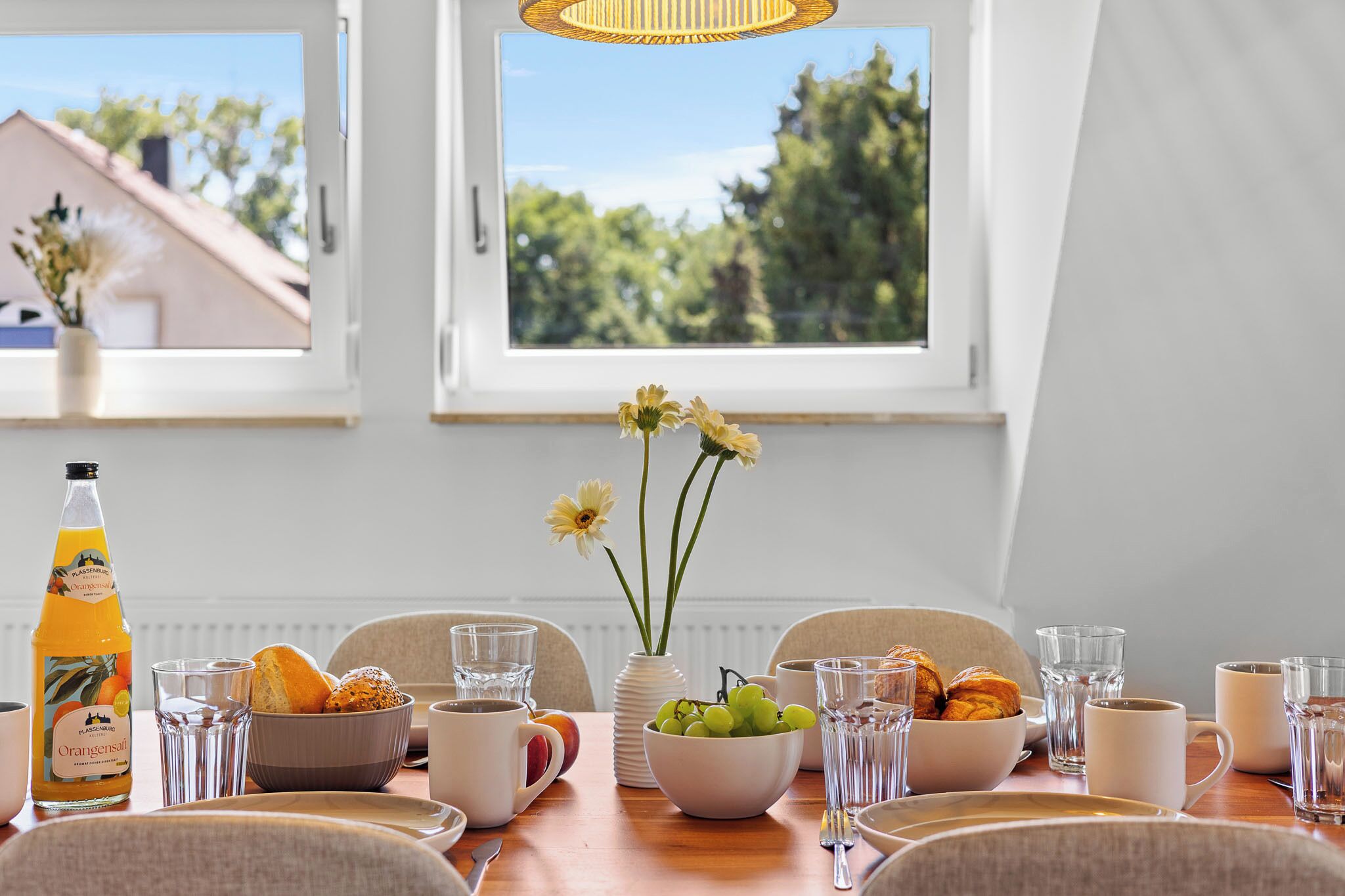 A breakfast table set with bread, pastries, a bottle of orange juice, a bowl of grapes, and a vase with flowers.