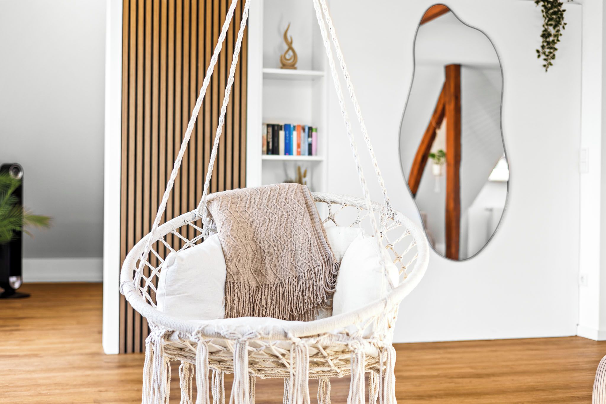 A macrame hanging chair with a cushion and a blanket, a wooden floor, a bookshelf, and a mirror on the wall.
