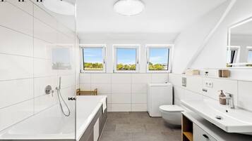 Une salle de bain moderne équipée d’une baignoire, de toilettes et d’un lavabo. Elle comporte trois fenêtres qui laissent entrer la lumière naturelle et offrent une vue sur l’extérieur.
