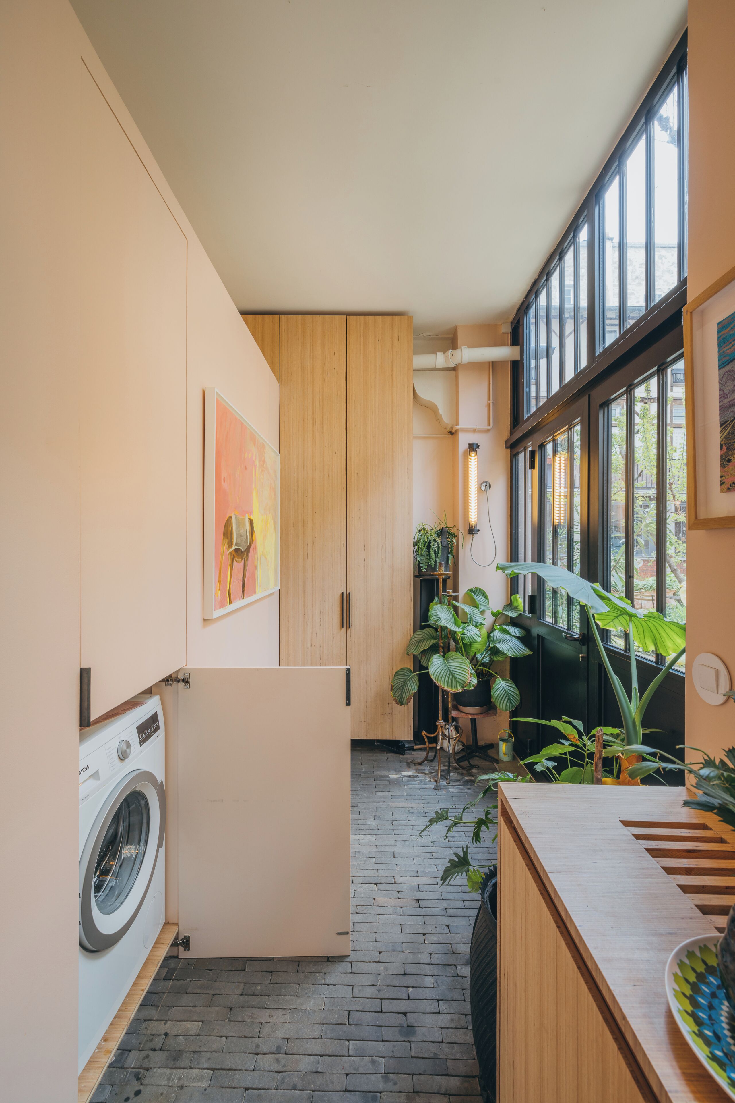 Une cuisine compacte équipée d’un lave-linge, de meubles en bois, d’une grande fenêtre et de plantes d’intérieur.