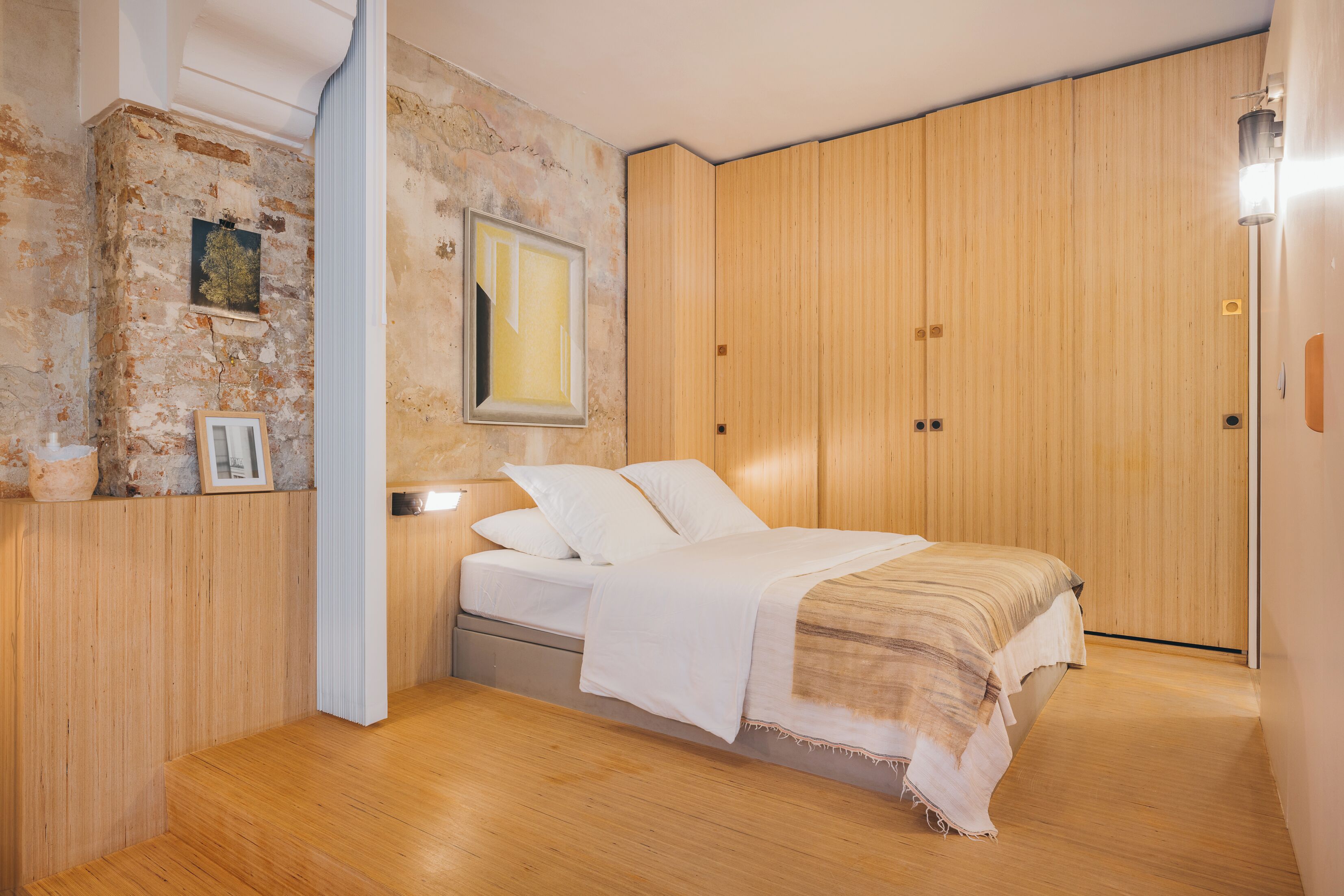 Une chambre à coucher avec un lit en bois, une table de chevet, une lampe fixée au mur et une armoire en bois.