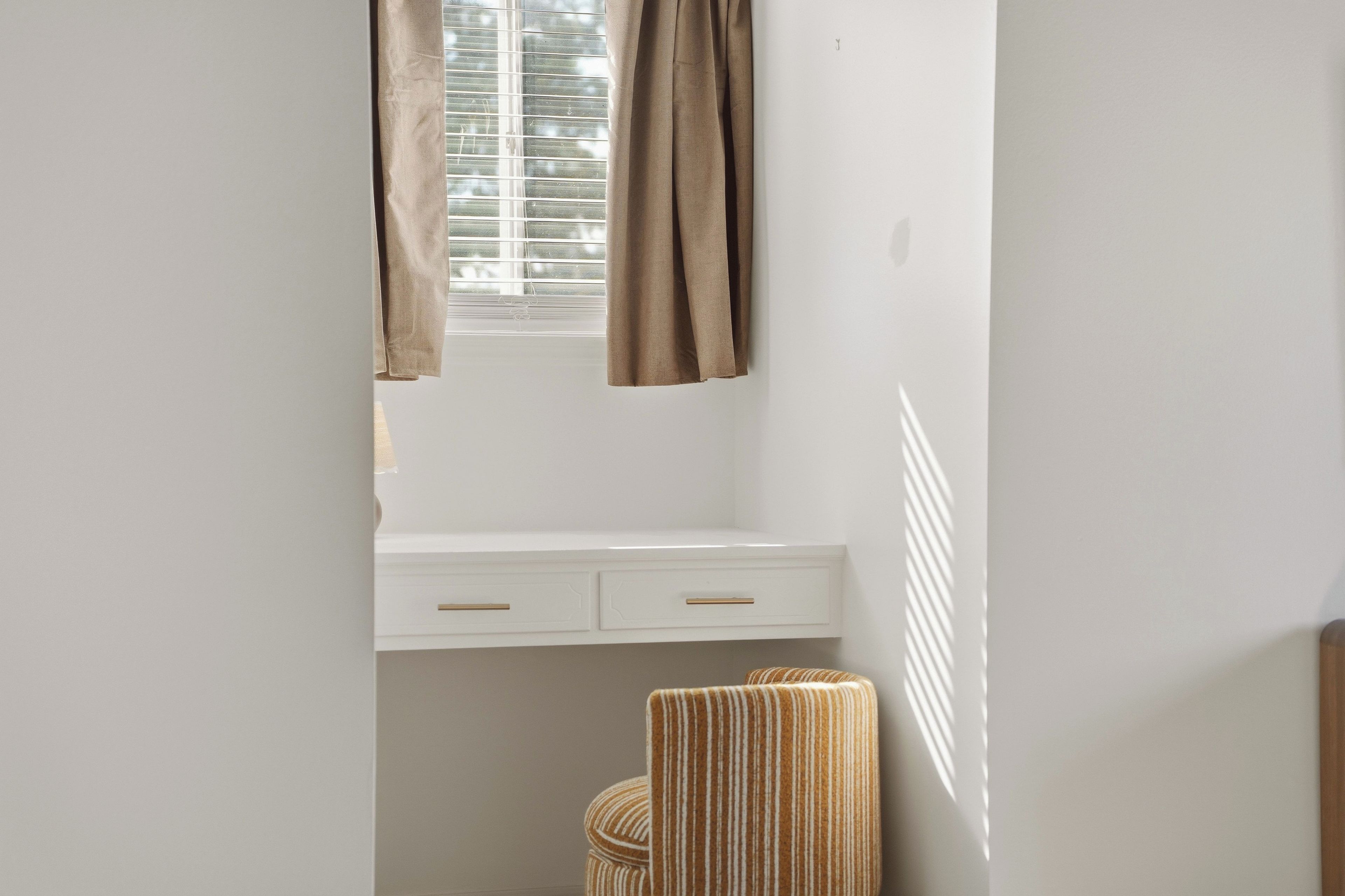 Una habitación con ventana, una mesita de noche blanca con cajones y una silla a rayas.