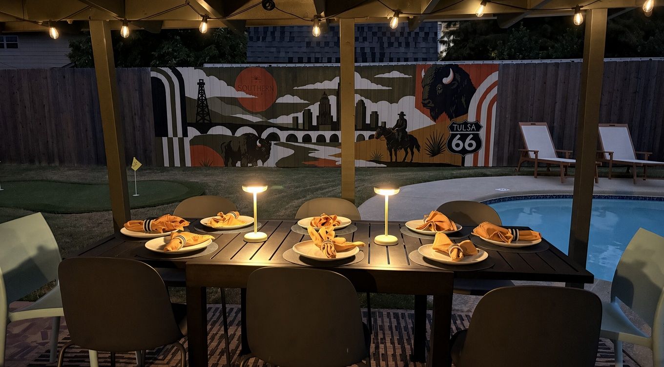Un área de comedor al aire libre con mesas dispuestas para cuatro, cada una con una vela y servilleta. Se observa un mural al fondo que representa un paisaje urbano y un bisonte.