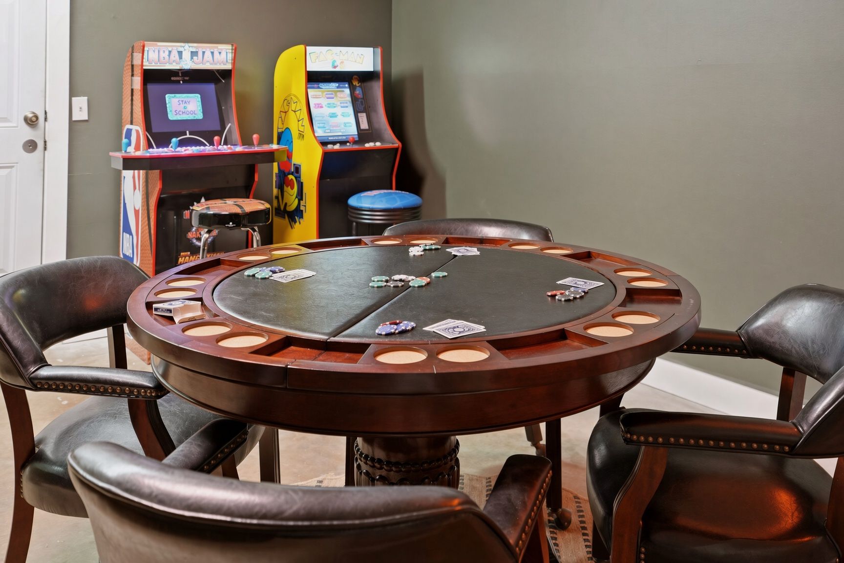A poker table with chips and cards, surrounded by leather chairs, in a room with arcade games in the background.