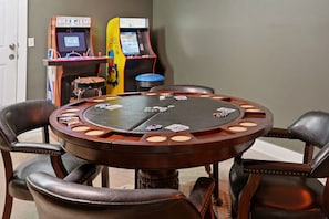 A poker table with chips and cards, surrounded by leather chairs, in a room with arcade games in the background.