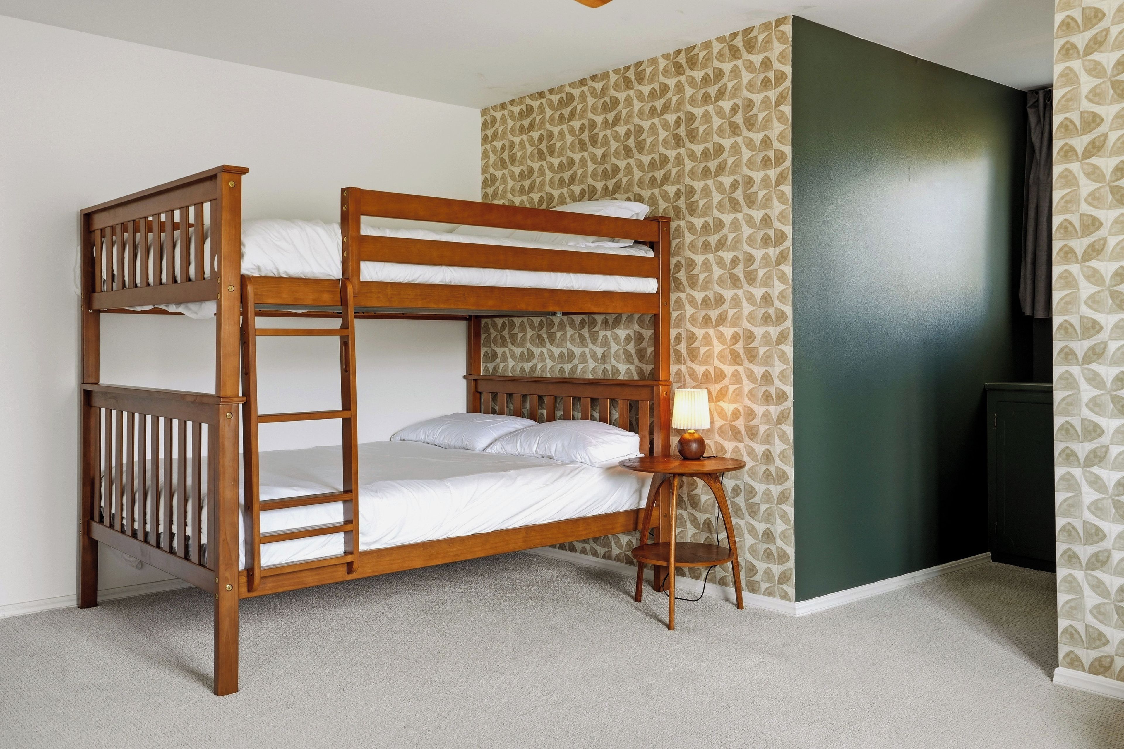 A bunk bed with a wooden frame, a small bedside table with a lamp, and patterned wallpaper.