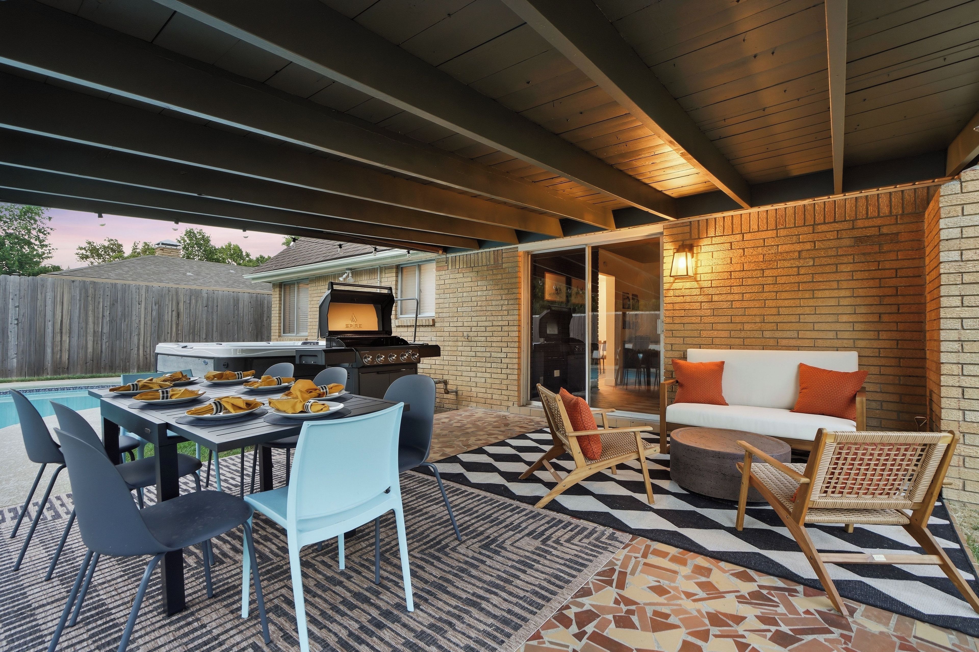 Un patio al aire libre con una mesa de comedor dispuesta para cuatro personas, rodeada de sillas, una parrilla y una zona de estar con un sofá y sillones.