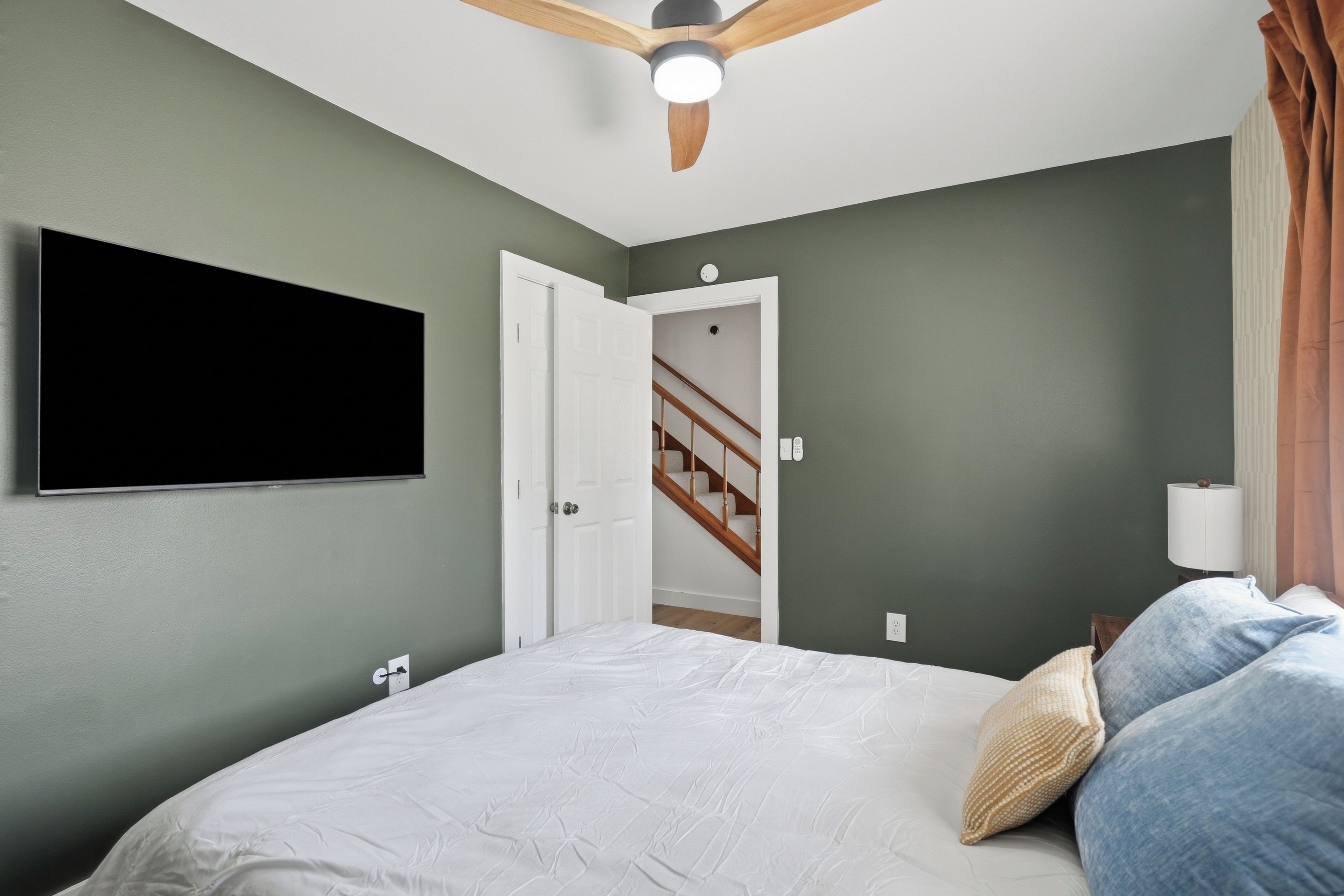 Un dormitorio con una cama, un ventilador de techo, un televisor montado en la pared y una puerta que da a una escalera.