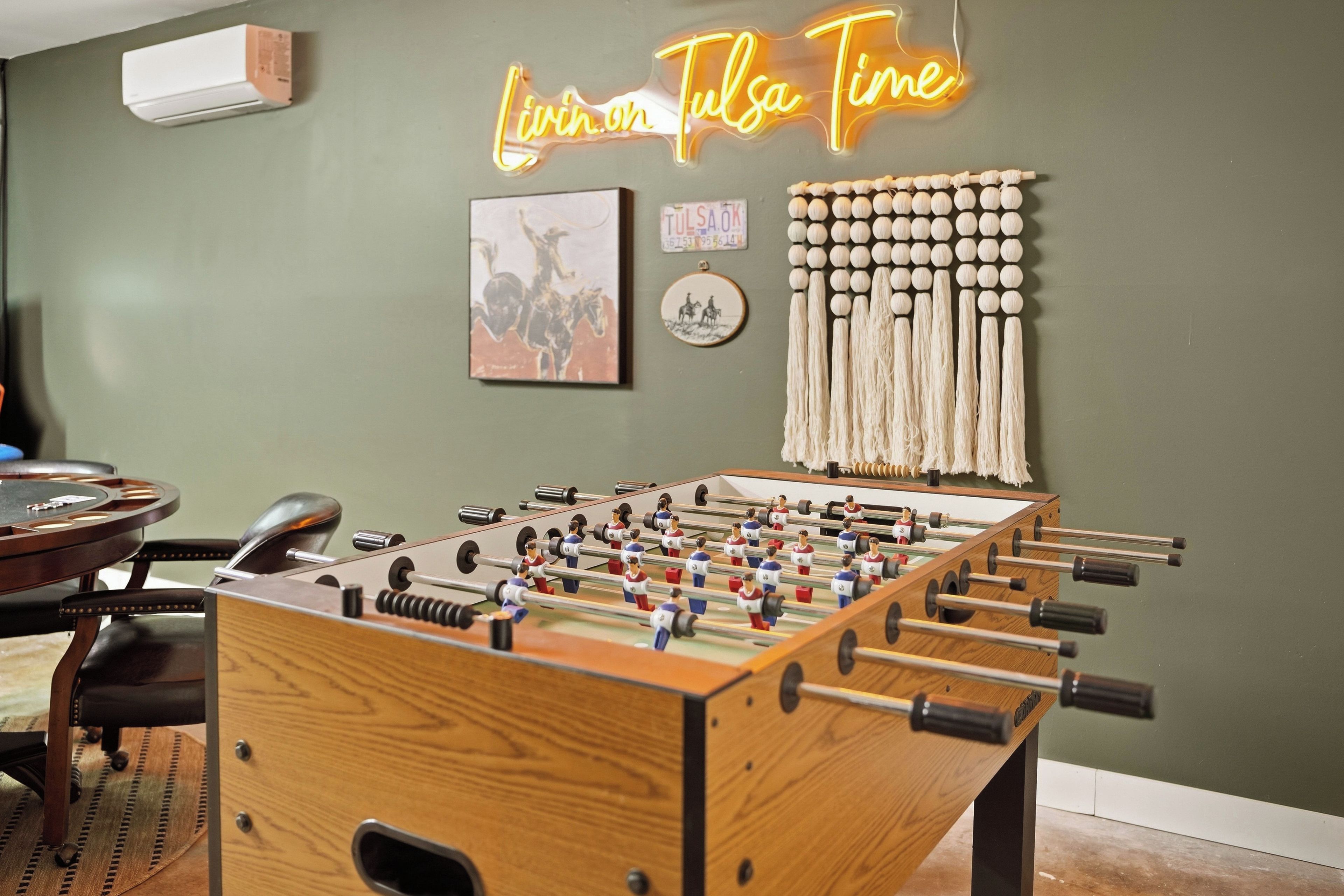 Una habitación con una mesa de futbolín, un letrero de neón que dice 'Living in Tulsa' y una pieza de macramé colgada en la pared.