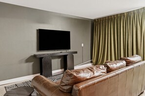 Ein Wohnzimmer mit einem Flachbildfernseher auf einem Lowboard, einem Ledersofa, einem gemusterten Teppich und einem grünen Vorhang.