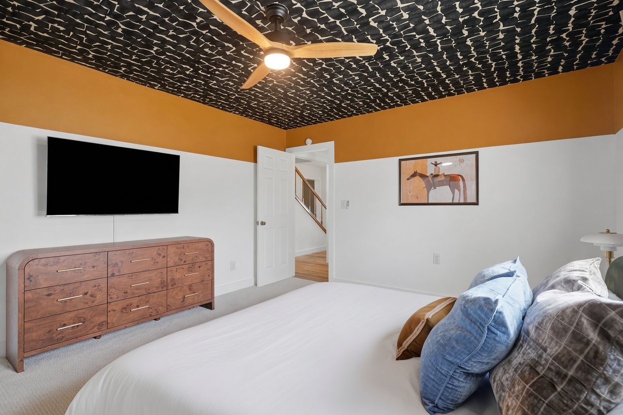 A bedroom with a bed, a wooden dresser, a flat-screen TV, a ceiling fan, a staircase, and a framed picture on the wall.