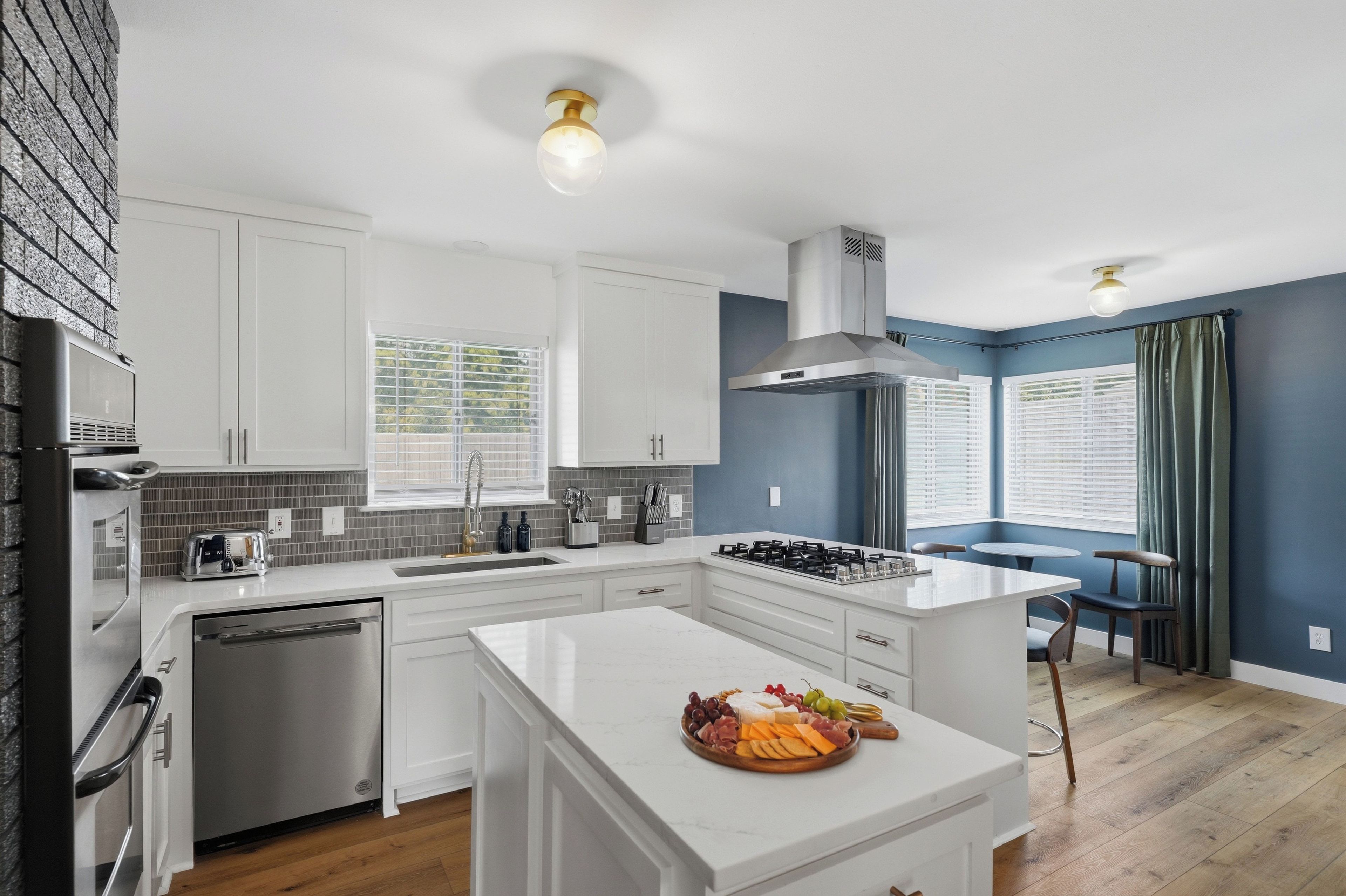 Una cocina moderna con armarios blancos, una isla central con una bandeja de frutas y quesos, lavavajillas integrado y una campana extractora de acero inoxidable.
