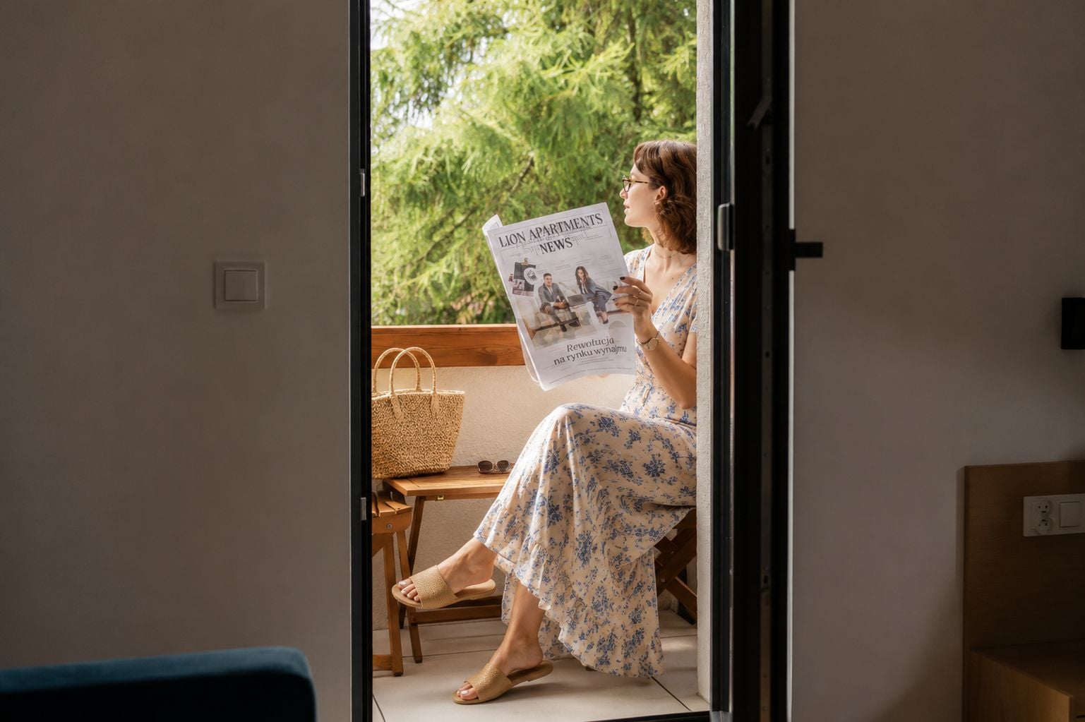 Eine Person sitzt am Fenster und liest eine Zeitung, während ein Korbwesen und ein Holzstuhl zu sehen sind.