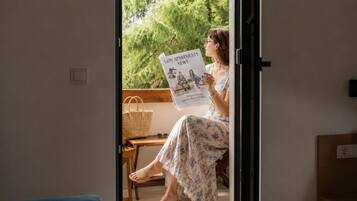 Eine Person sitzt am Fenster und liest eine Zeitung, während ein Korbwesen und ein Holzstuhl zu sehen sind.