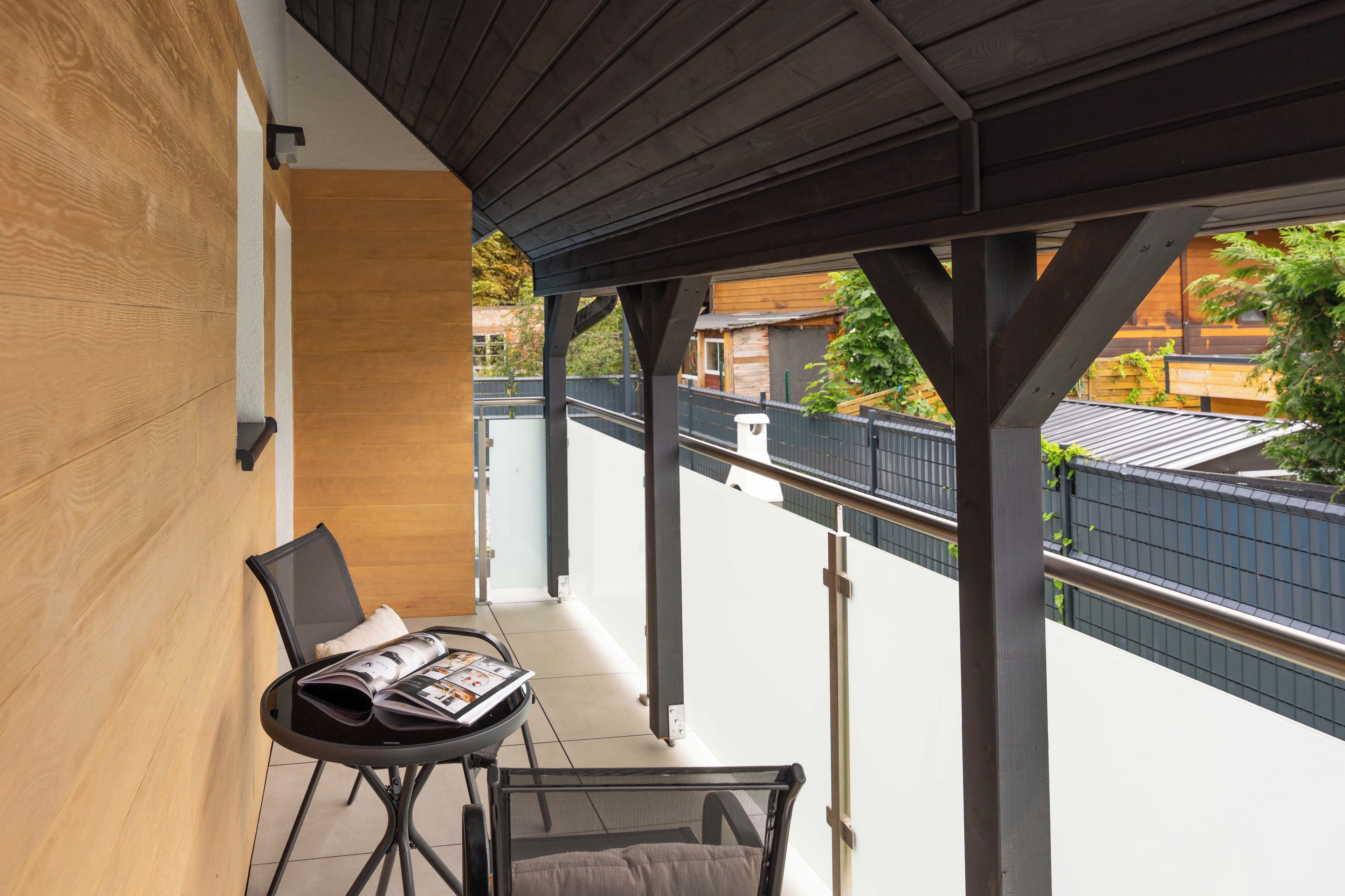 Ein überdachter Balkon mit Tisch und Stühlen, eine Holzverkleidung und Blick auf ein Gebäude, das durch einen Zaun abgetrennt ist.