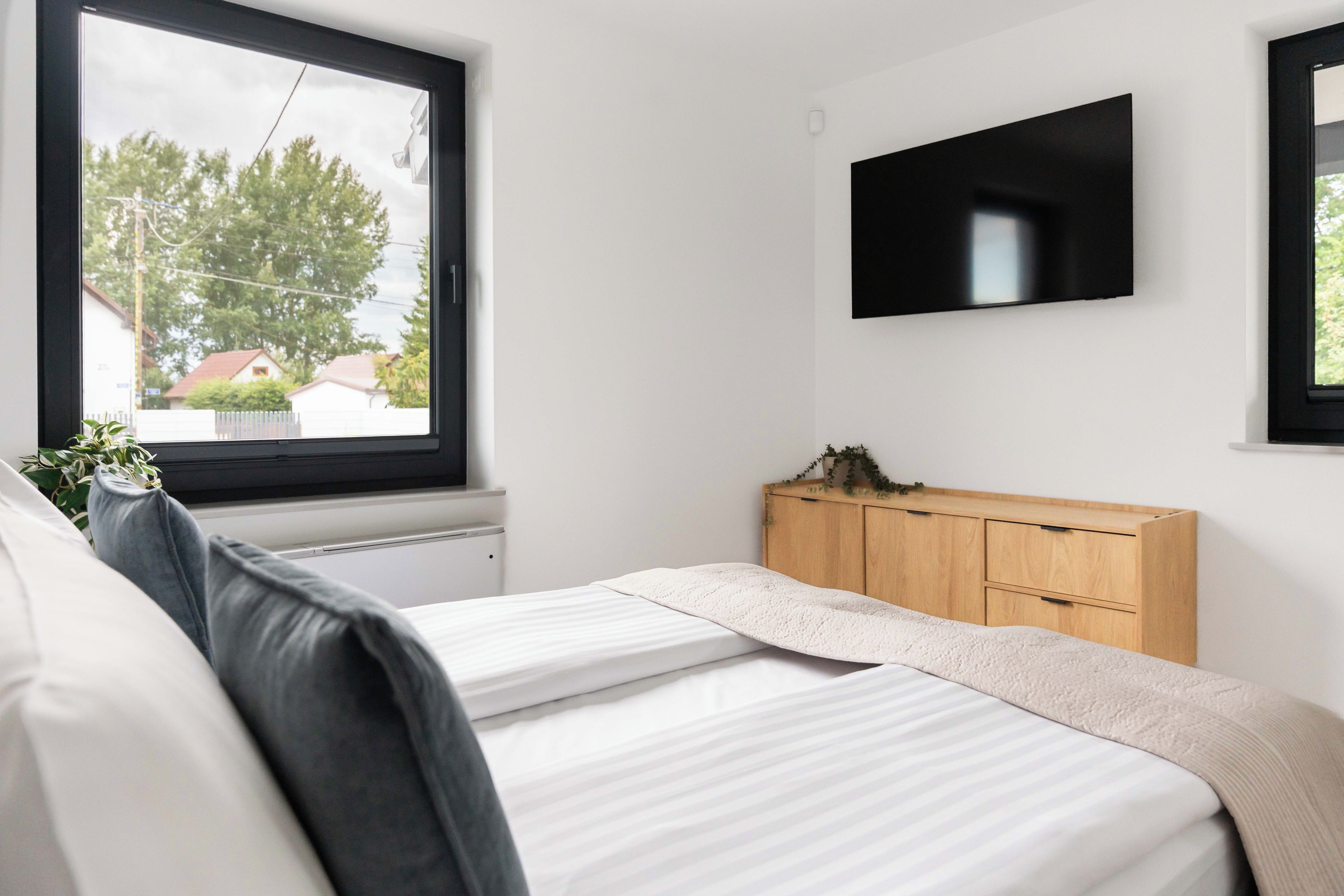 Ein Schlafzimmer mit einem Bett, einem an der Wand befestigten Fernseher, einem Fenster mit Blick auf Häuser und einem Holzkoffer.