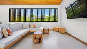 Un soggiorno moderno con soffitto in legno, un'ampia finestra vista su del verde, un divano grigio con cuscini decorati e un tavolino rotondo in legno.
