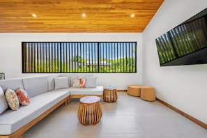 A modern living room with a wooden ceiling, a large window with a view of greenery, a grey sofa with patterned cushions, and a round wooden table. - Tamarindo Compound w/ Pools & Stunning Views (Tamarindo)