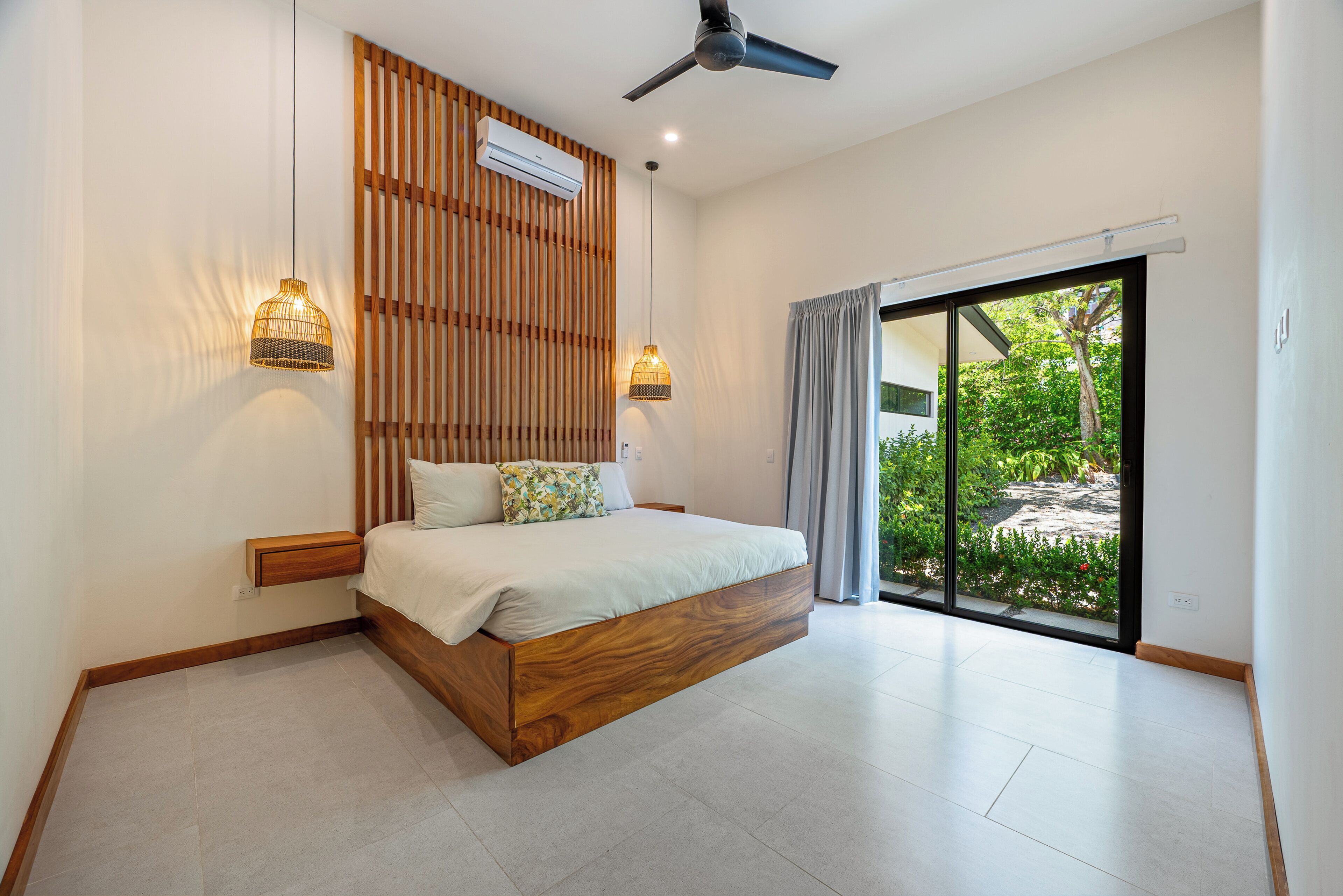 A modern bedroom with a wooden bed frame, a ceiling fan, and a view of a garden through a sliding glass door.