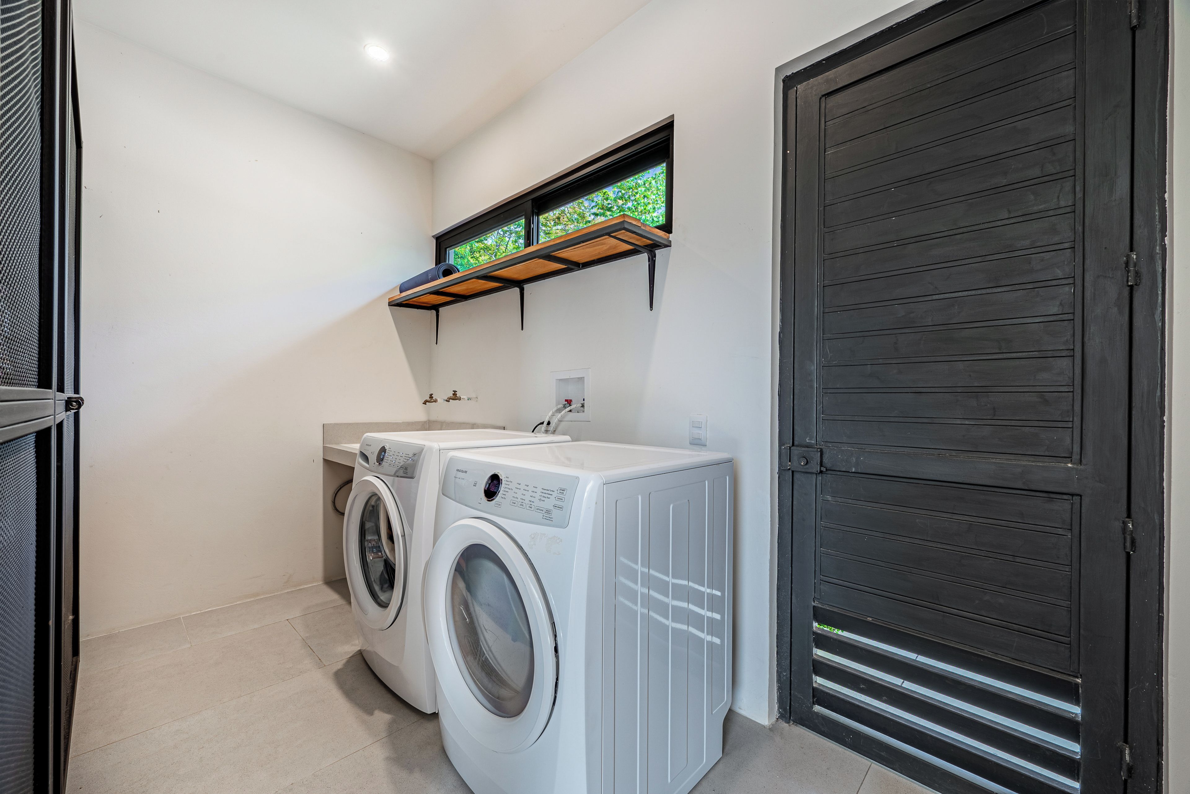 A laundry room with two front-loading washing machines, a small window with a view of greenery, and a dark wooden door.