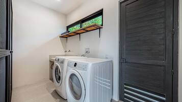 A laundry room with two front-loading washing machines, a small window with a view of greenery, and a dark wooden door.