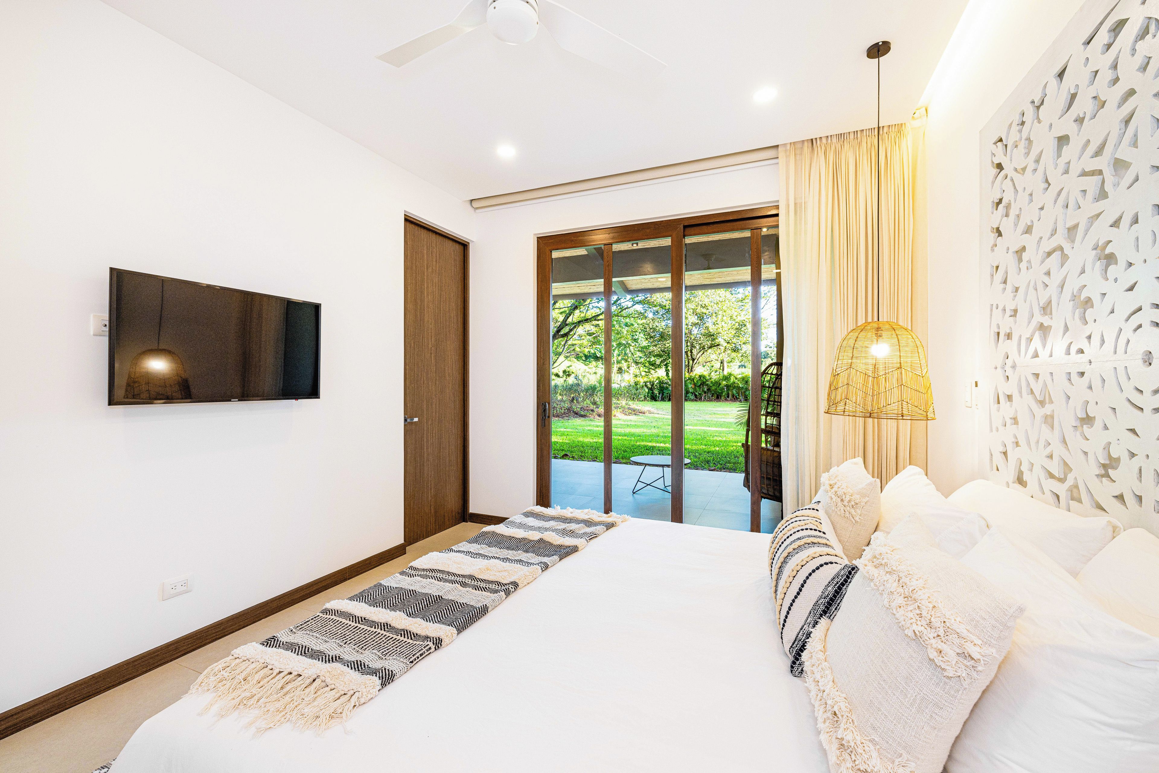 Une chambre à coucher comprenant un lit, un téléviseur, un ventilateur de plafond et une vue sur un jardin, accessible par une porte coulissante.