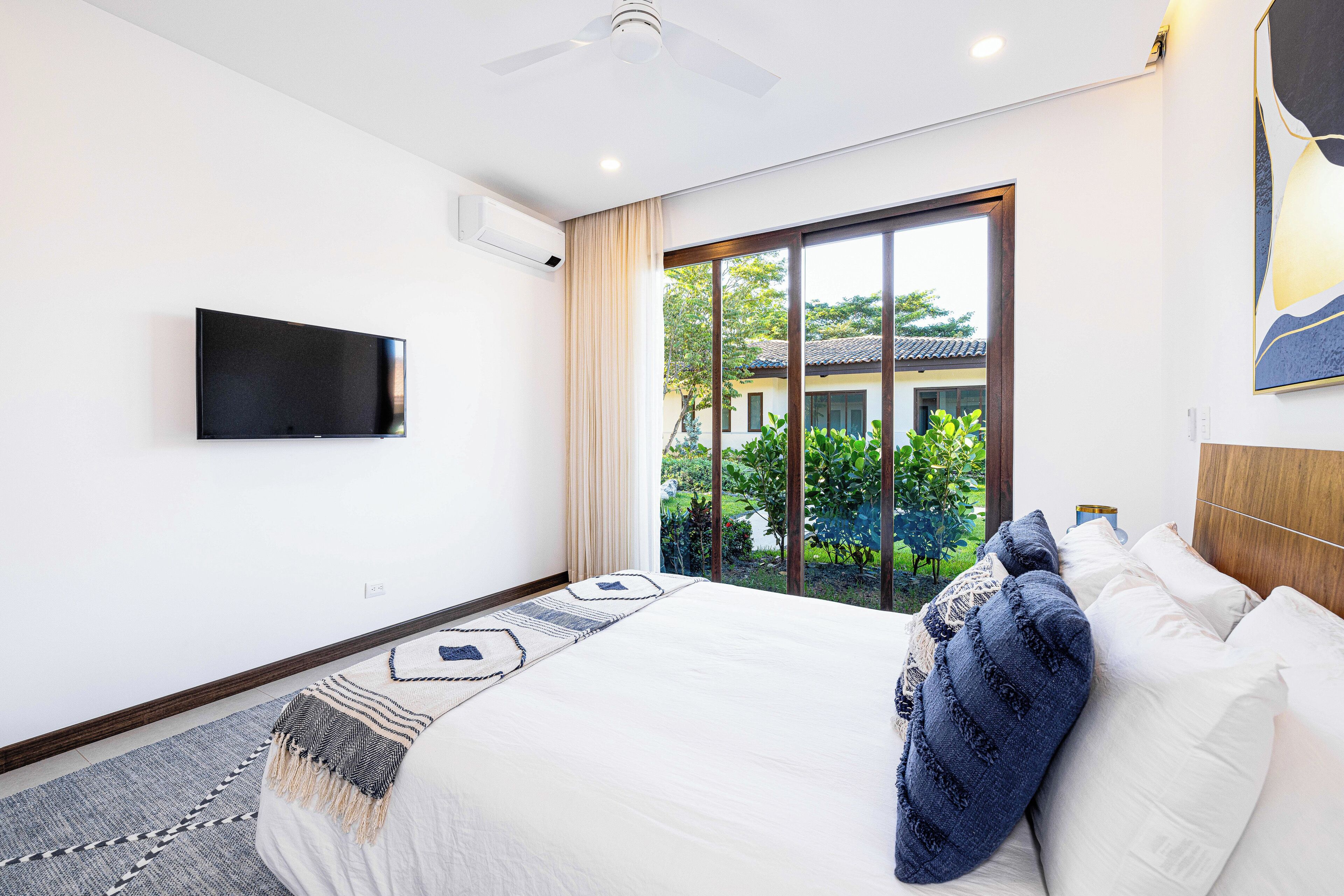 A modern bedroom with a large bed, a flat-screen TV mounted on the wall, and a view of greenery outside.