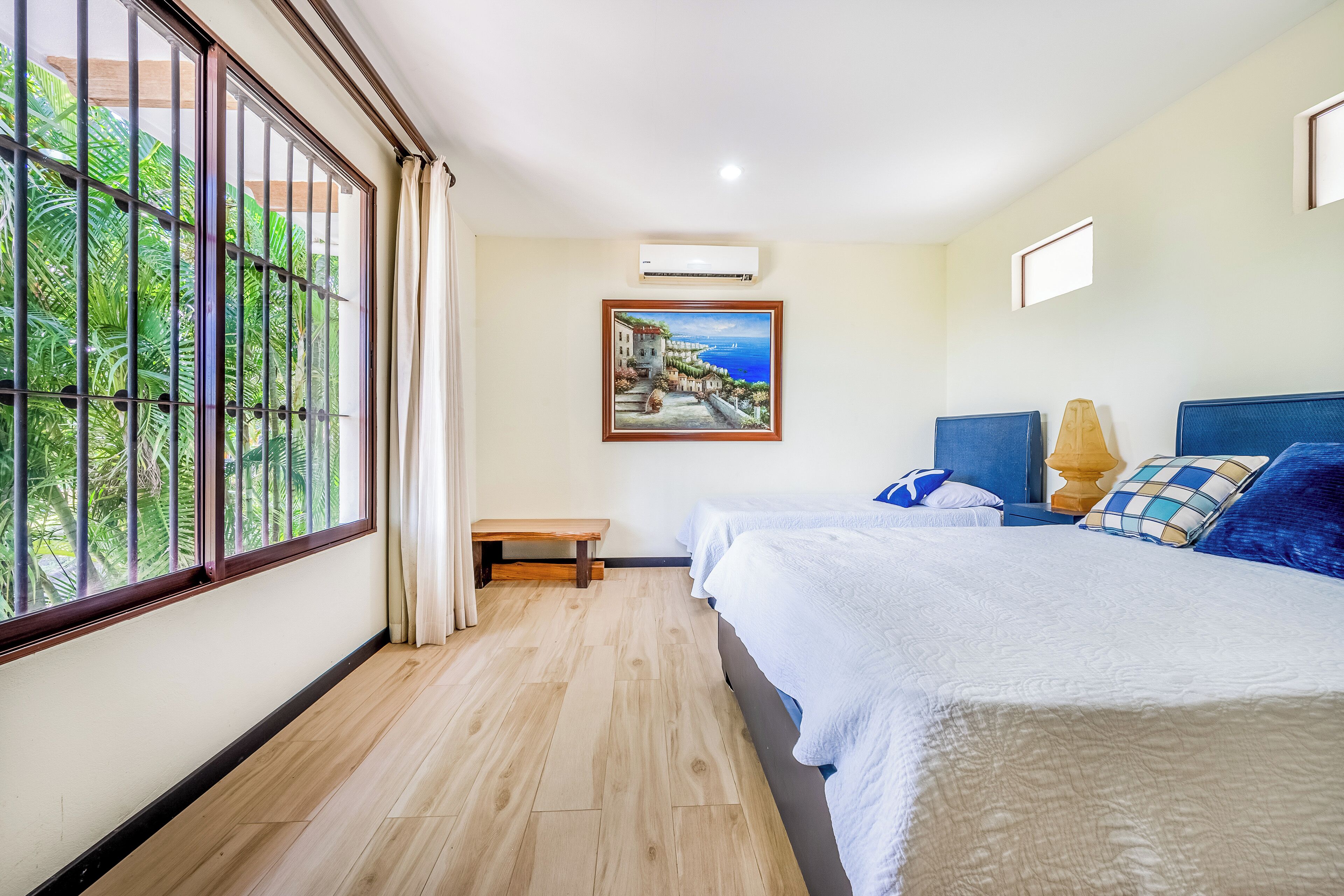 A hotel room with a large window, a painting, a bench, and a bed with pillows.