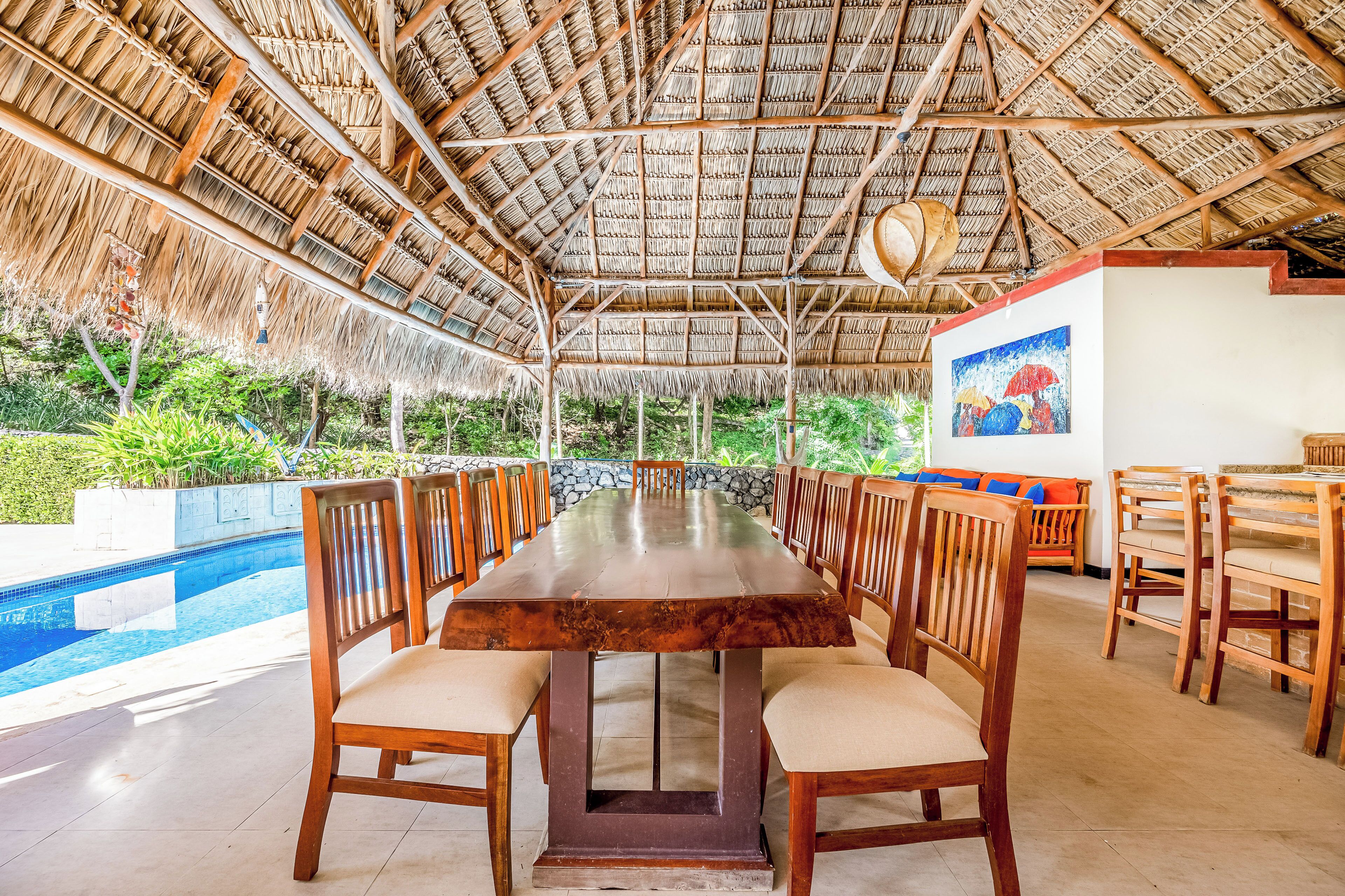 A long dining table with chairs, a pool, and a thatched roof structure.